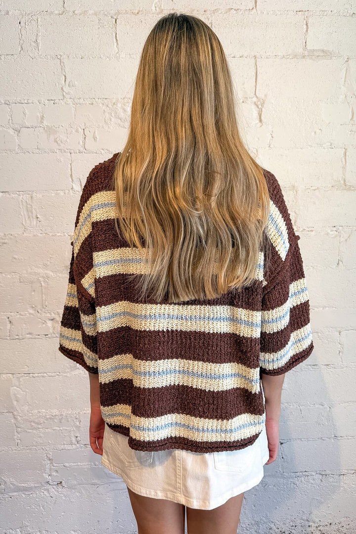 A woman viewed from behind wearing a brown, cream, and light blue striped knit cardigan sweater, standing against a white brick wall.