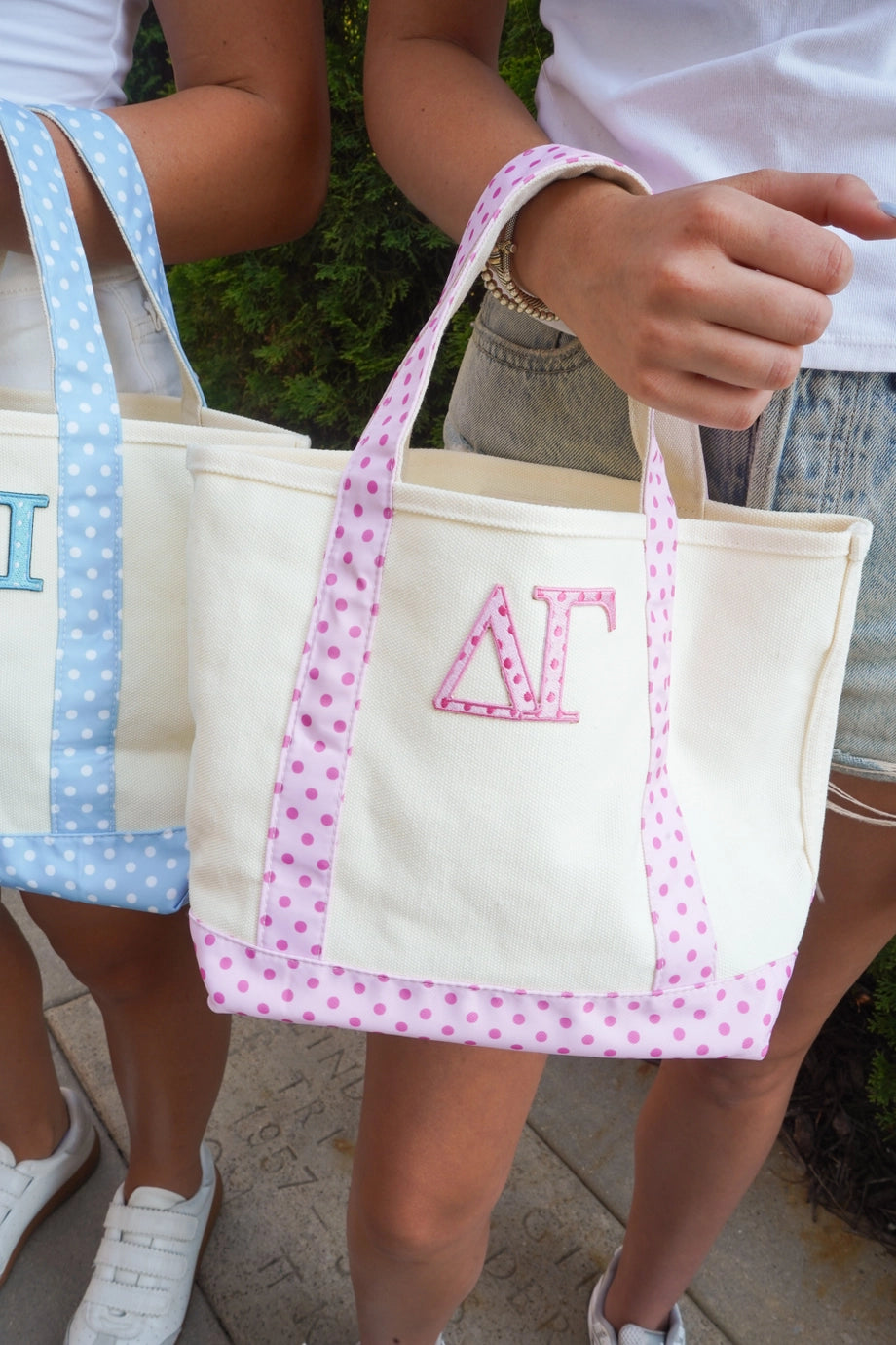 A cream canvas tote bag with pink polka-dot handles and bottom trim, featuring an embroidered pink Greek letter Delta Gamma monogram, held by a person outdoors.