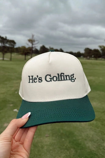 Cap with 'He's Golfing' text held in front of a golf course