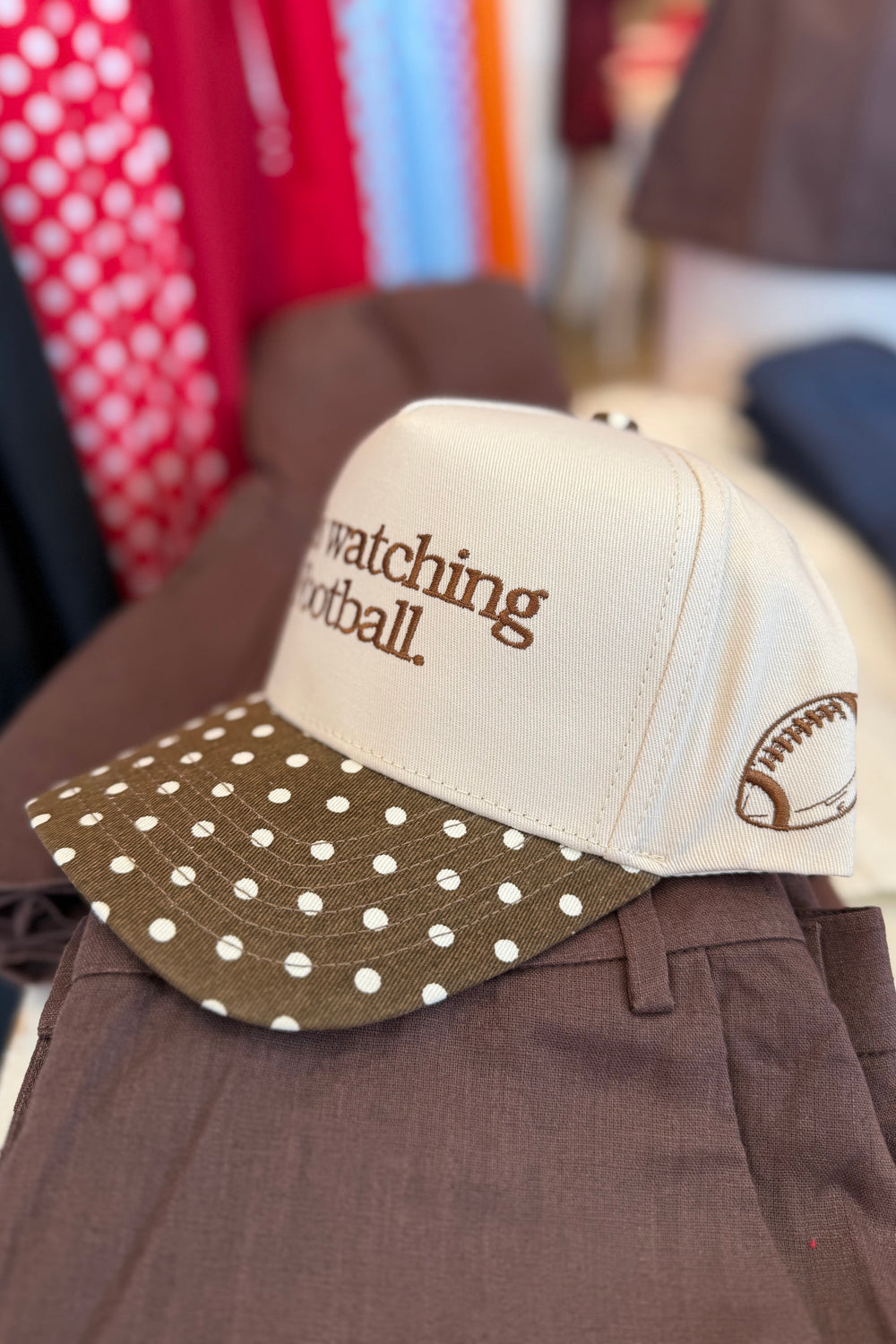 A cream and brown baseball cap with 'watching football.' embroidered on the front, resting on brown pants in a clothing store setting.