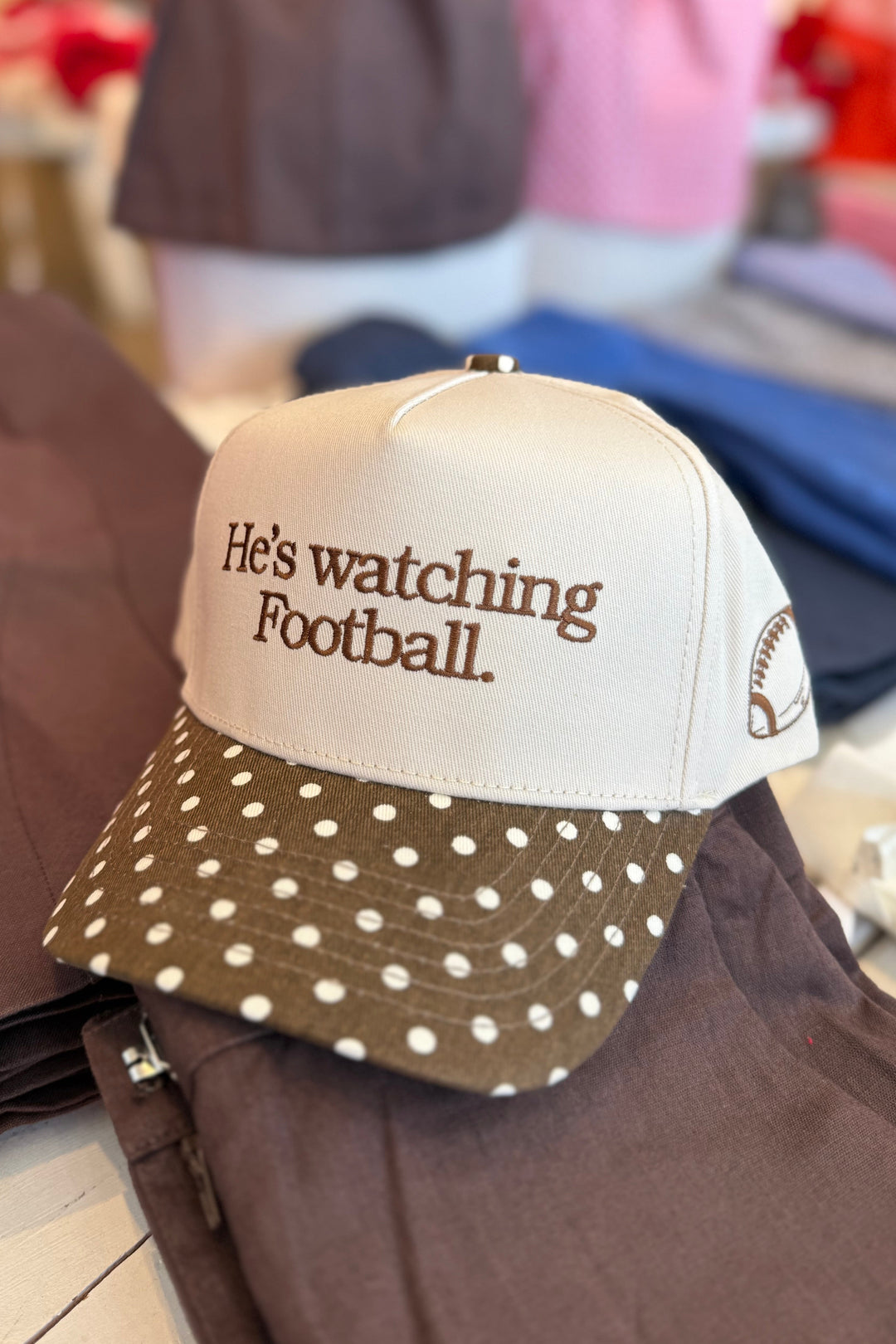 A baseball cap with a cream crown and a brown polka-dot visor, featuring the embroidered text 'He's watching Football.' on the front.