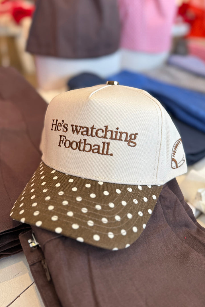A baseball cap with a cream crown and a brown polka-dot visor, featuring the embroidered text 'He's watching Football.' on the front.