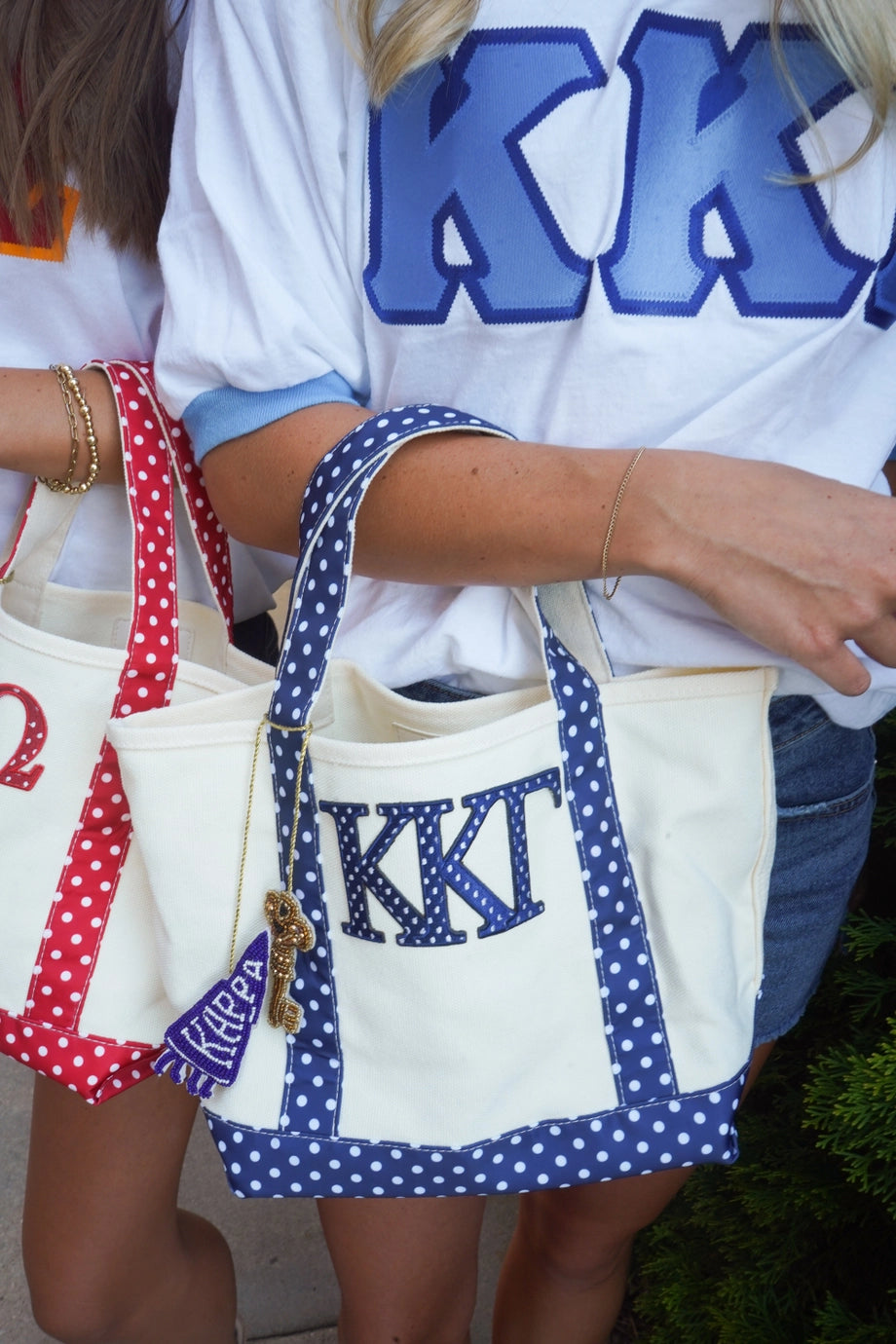 Two cream canvas tote bags with polka-dot trim and monogrammed 'KKT' lettering, carried by people wearing white 'KK' shirts. One bag has red polka-dot trim and handles, and the other has blue polka-dot trim and handles.