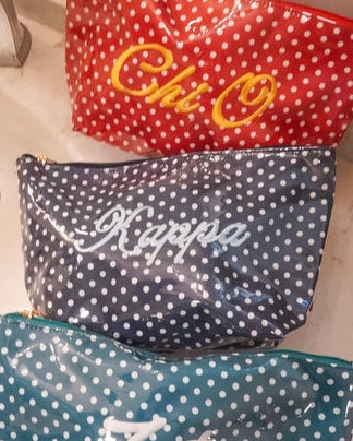 Three personalized cosmetic pouches with white polka dots are lined up on a bathroom sink. The top red pouch is embroidered with 'Chi Omega' in yellow, the middle navy blue pouch is embroidered with 'Kappa' in white, and the bottom teal blue pouch is embroidered with 'Zeta' in white.