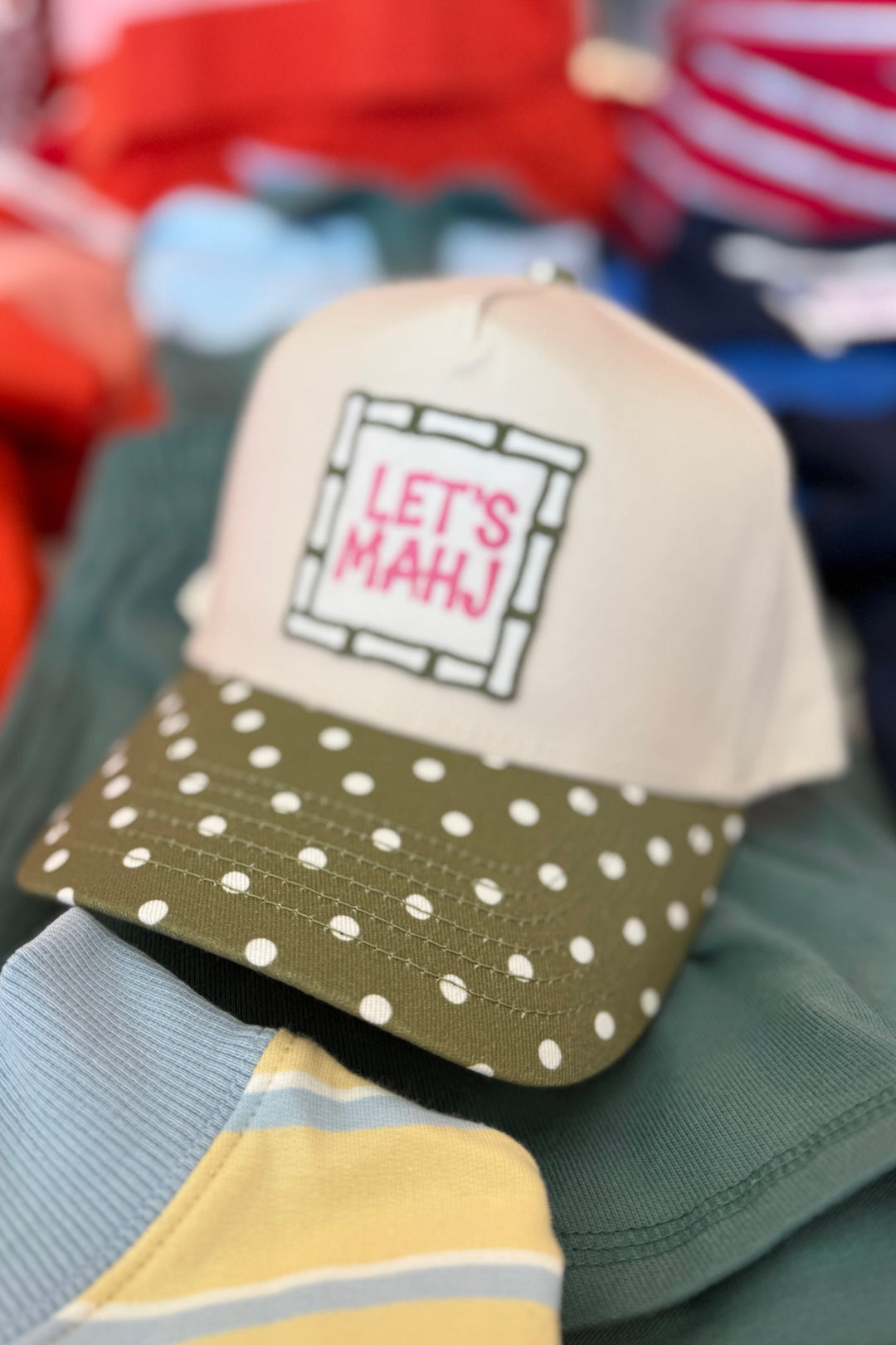 A beige and olive green trucker hat with white polka dots on the brim and a patch reading 'LET'S MAHJ', resting on a pile of colorful clothing.