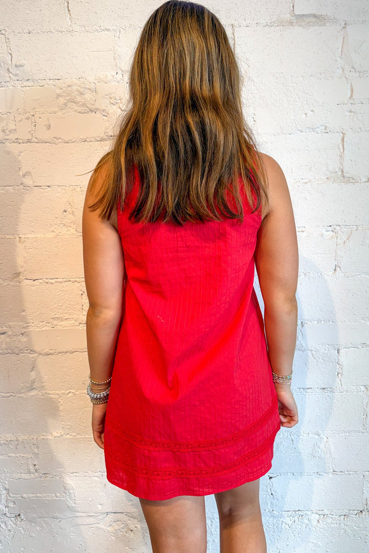A woman viewed from behind wearing a sleeveless red dress, standing against a white brick wall.