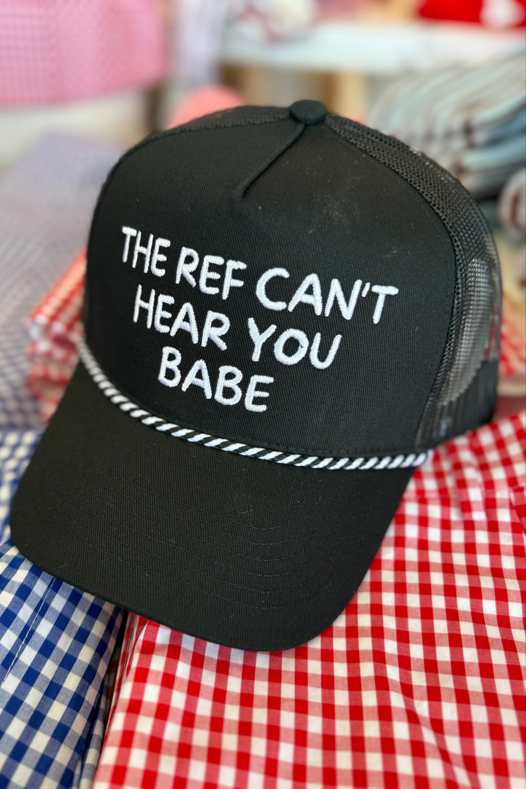 A black trucker-style cap with white embroidered text reading 'THE REF CAN'T HEAR YOU BABE' and a white rope-like embroidered detail across the front, displayed on red and blue gingham fabric.
