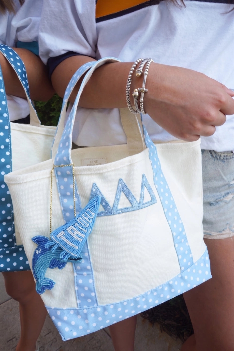 A close-up lifestyle shot of a person holding a cream canvas tote bag with light blue and white polka dot straps and trim, featuring a blue embroidered fish charm and geometric triangle pattern.