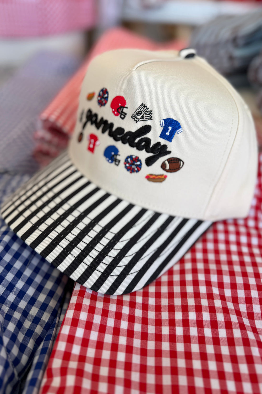 A cream-colored baseball cap with a black and white striped visor, featuring the embroidered text 'Pamedoy' and various sports-themed patches. The cap is styled on top of red, white, and blue gingham shirts.