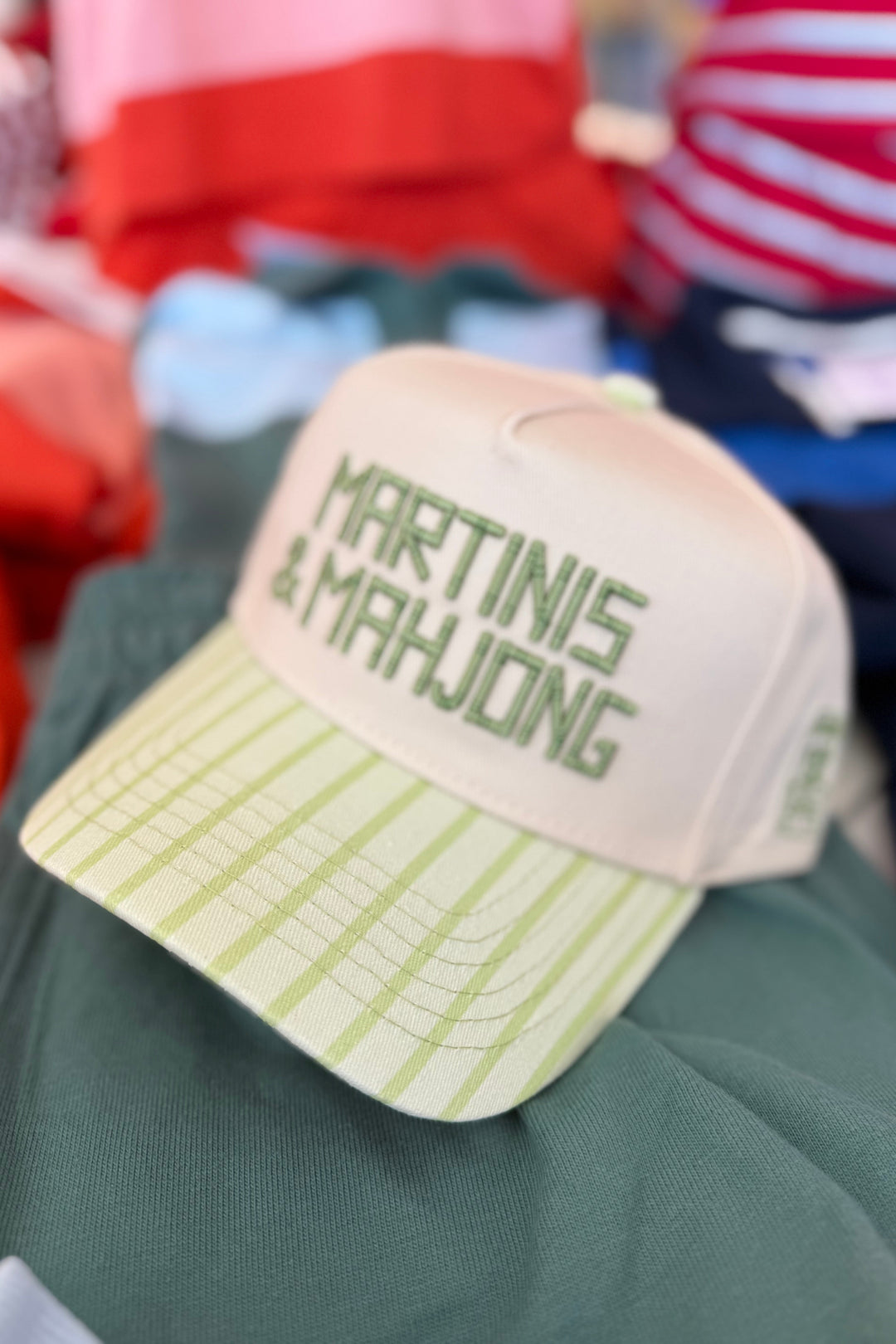 A baseball cap with a cream crown and a light green plaid brim, featuring the embroidered text 'MARTINIS & MAHJONG' in green on the front. The cap is resting on a dark green fabric surface with other colorful clothing items blurred in the background.