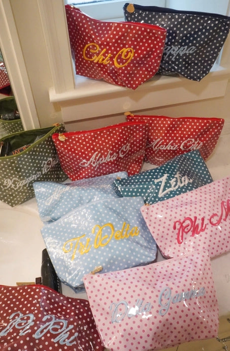 A group of embroidered zippered pouches in red, blue, green, and pink, all featuring white polka dots and white script lettering, displayed on a light-colored surface.